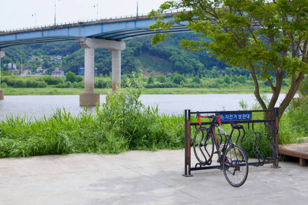 Gongju City, South Korea - May 27th, 2021: A road bike rests on a metal bike rack near a bridge spanning the Geum River, offering cyclists a resting point along the scenic cycling route.
