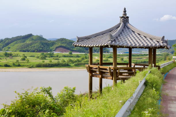 Gongju, South Korea - May 27th, 2021: A wooden pavilion with a tiled roof stands on a hillside along the Geum River, providing a scenic rest stop along the bike path with views of lush green fields.