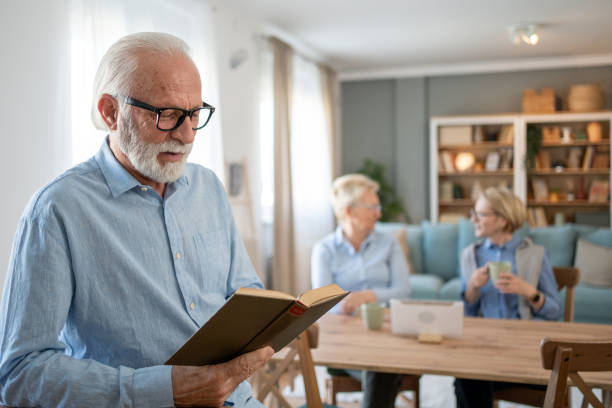 istock Senior man reading a book while two senior women chatting in the background 2202675332