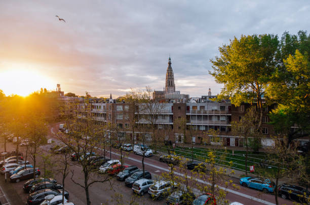 A breathtaking sunset view over Breda, Netherlands, featuring the iconic Gothic tower of Grote Kerk (Church of Our Lady) in the background. The golden sunlight casts a warm glow over the cityscape, illuminating the residential buildings and tree-lined streets. A seagull soars above, adding to the serene ambiance of the evening scene. The contrast between modern urban life and historical architecture captures the charm of this Dutch city