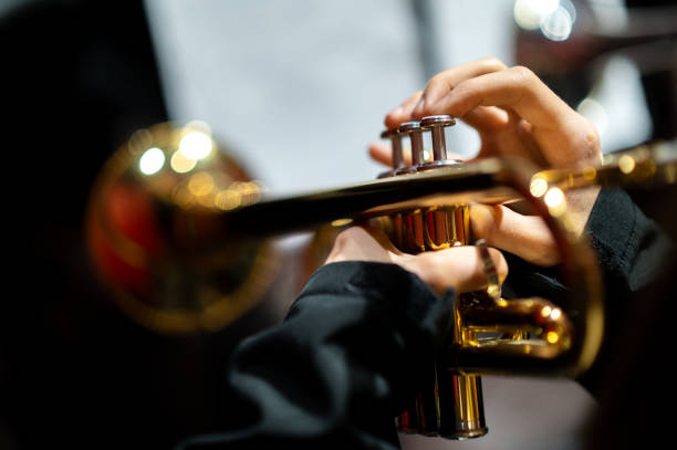 This close-up photograph captures the hands of a musician playing a trumpet, with a focus on the intricate details of the instrument's valves and the player's fingers pressing them. The golden brass surface of the trumpet reflects warm light, while the blurred background creates a bokeh effect, emphasizing the musician's skill and precision. The composition highlights the artistry and dedication behind playing a brass instrument in a concert setting.