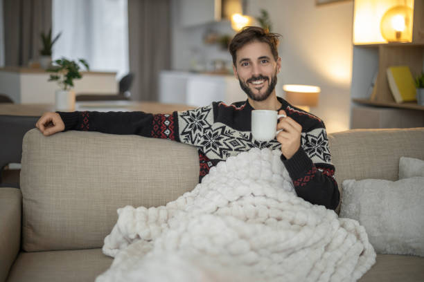 Man enjoying a cozy moment with a warm drink while wrapped in a blanket on a comfortable sofa in a modern living room