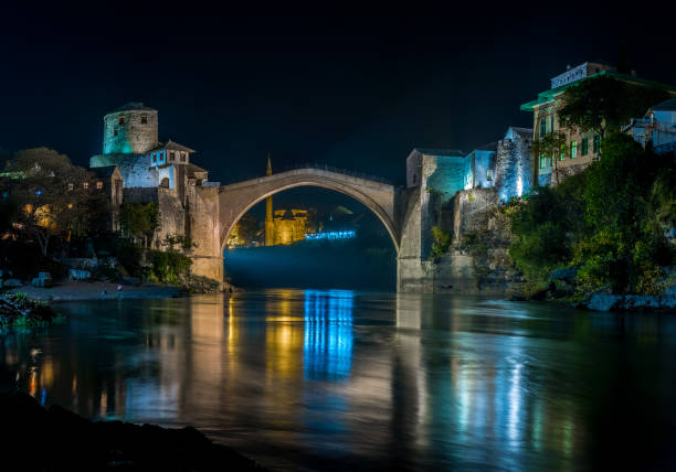 A stunning arched bridge is illuminated at night, reflected in the river below. Buildings on either side add to the architectural display.