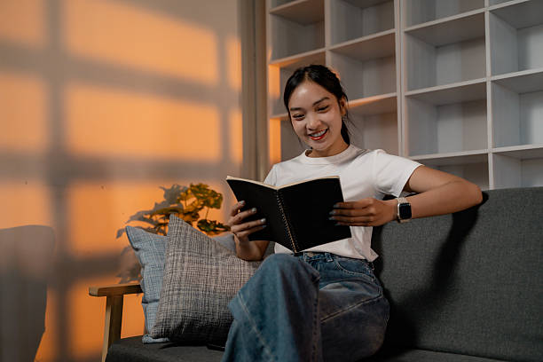 istock Young woman reading a book sitting on a sofa at home 2202686942