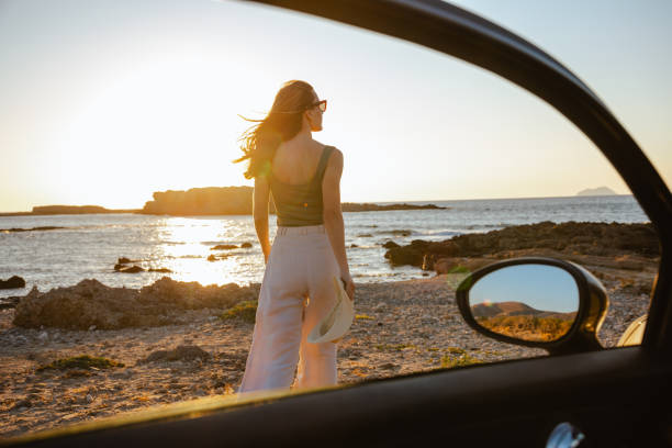 A woman stands on a rocky beach at sunset, appreciating the peaceful coastal scenery. The beautiful seaside ambiance and the setting sun create a captivating and calming atmosphere.