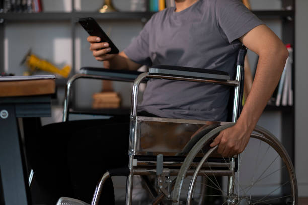istock Young man sitting in wheelchair using smartphone at home office 2202708734