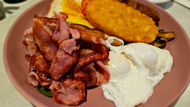istock A hearty breakfast plate featuring crispy bacon, poached eggs, hash browns, toast, sausage, and sautéed mushrooms on a pink plate. The meal is rich and indulgent, perfect for a filling start to the day. 2202729465
