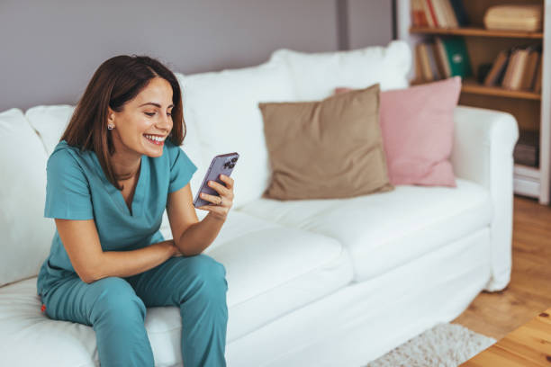 istock Smiling Woman in Scrubs Using Smartphone on Comfortable White Sofa 2202738489