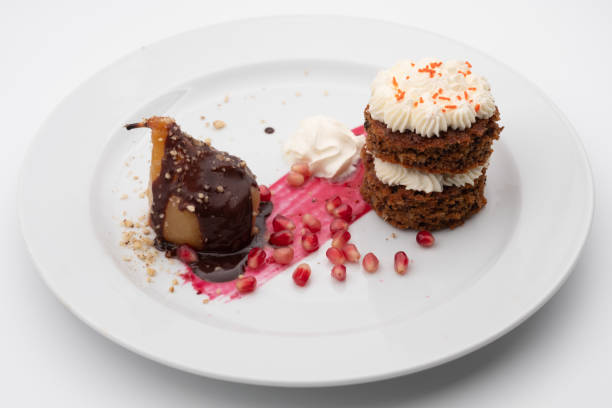 Elegant plated dessert features chocolate covered pear carrot cake whipped cream and pomegranate seeds on a white plate A sophisticated dessert on a white plate. Features a pear covered in chocolate and nuts, a stack of carrot cake with cream, whipped cream, and pomegranate seeds for garnish. Set against a plain backdrop. plated chocolate dessert stock pictures, royalty-free photos & images