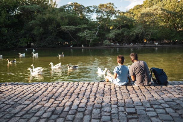 A tranquil scene of a father and son sitting near a lake feeding ducks, surrounded by nature's beauty, illustrating family bonding and peaceful outdoor moments in harmony with the environment.