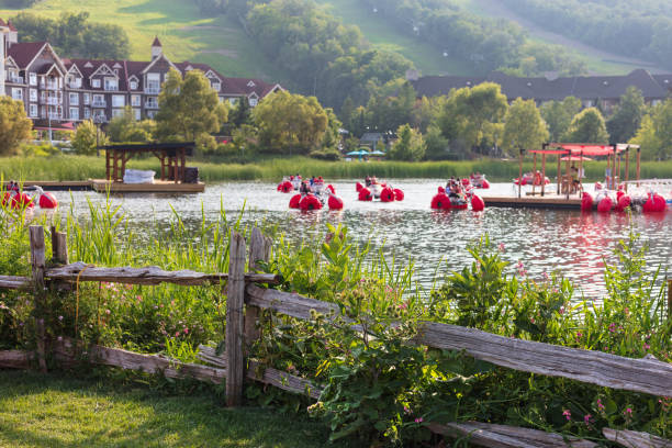 The Blue Mountains, Ontario, Canada - August 2, 2023: Village cabins with mountain in the background on a sunny summer day. Lake in the foreground with people in paddle boats.