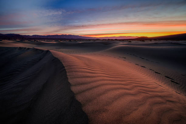 Golden light casts intricate patterns on the rippled sand of the Mesquite Flat Sand Dunes in Death Valley National Park, as the sun rise behind distant mountain peaks. The delicate interplay of light and shadow enhances the undulating curves of the dunes, creating a sense of depth and movement. A few scattered footprints hint at the vastness and solitude of this harsh yet stunning desert landscape. The vivid hues of the sky contrast beautifully with the earthy tones, offering a mesmerizing scene of nature’s artistry.