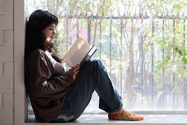 istock female student sitting by the window reading a book 2202760328