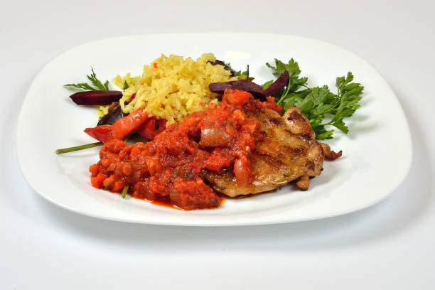 A delicious grilled chicken breast is served on a black plate alongside bright red chili peppers and a colorful mix of onions and herbs. A portion of fluffy rice completes the meal.