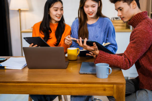 istock Three Asian young adult students sit around the kitchen table, attending online class on tablet. They engage in video conferencing, discussing lessons, taking notes, digital learning experience. 2202777153