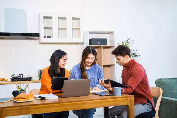 istock Three Asian young adult students sit around the kitchen table, attending online class on tablet. They engage in video conferencing, discussing lessons, taking notes, digital learning experience. 2202777154