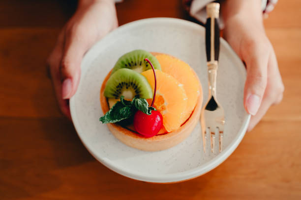 A Fresh Fruit Tart on White Plate with Cherry and Mint Garnish Held by Hands Over Wooden Table Background