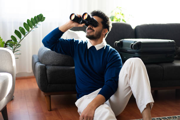 Young man sitting on the floor using binoculars, dreaming of his next trip with his suitcase next to him