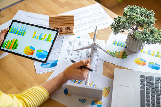 istock Businesswoman analyzing data on a tablet, showcasing charts and graphs alongside a wind turbine model and miniature house, emphasizing investments in renewable energy 2202790877