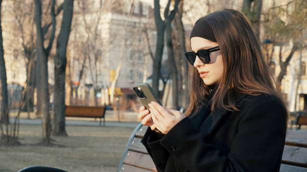 istock Young woman using smartphone sitting on bench in a park 2202794784