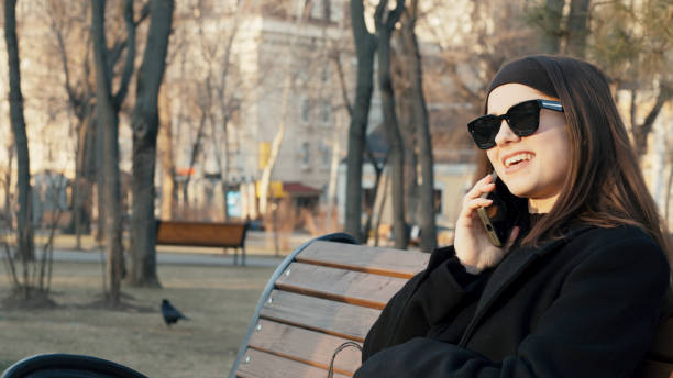 istock Young woman talking on phone and smiling while sitting on bench in park 2202794789