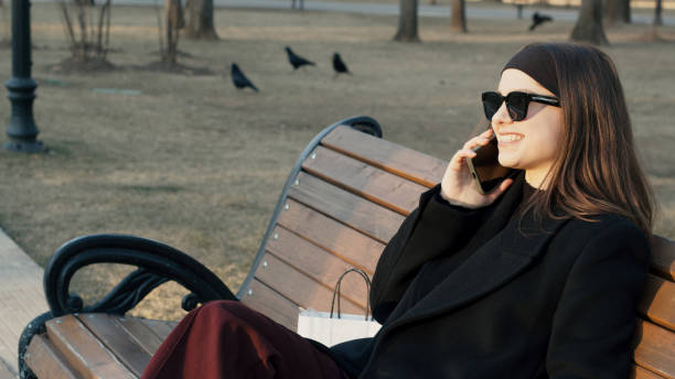 istock Young woman talking on smartphone and smiling while sitting on bench in park 2202794794