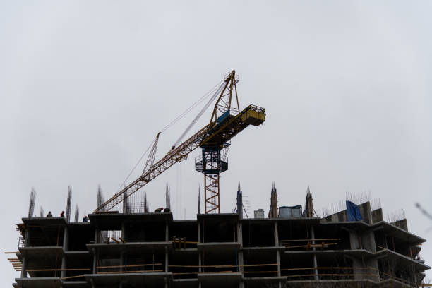 Russia, Podolsk - November 14, 2024: Construction crane is positioned at building site, actively lifting materials. Workers can be seen on-site as the grey clouds loom overhead, indicating an overcast day.