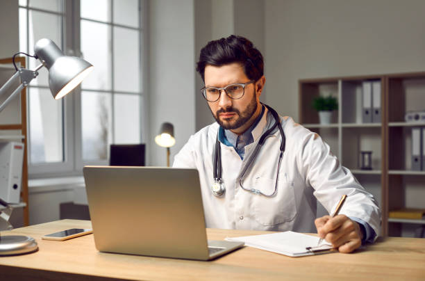 istock Male doctor sitting in office, working on laptop computer, doing research and taking notes 2202810391