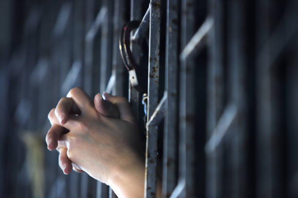 istock Hands Gripping Steel Bars in a Dimly Lit Prison Cell Setting 2202816486