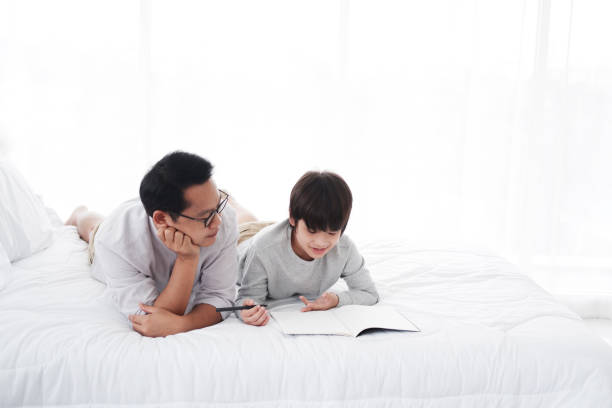 istock Smiling Asian father teaching homework with boy son for write with pencil on a notebook together on bed in bedroom at home. Family love and child educational for homeschool on holiday concept 2202818992
