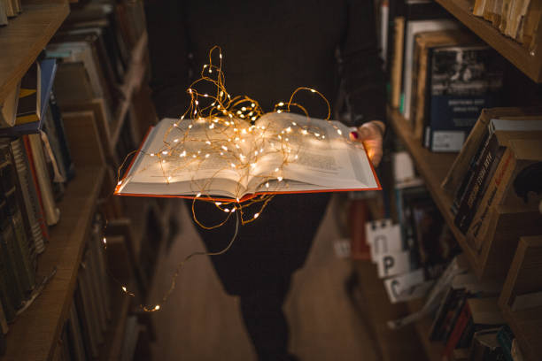 Unrecognizable librarian standing between bookshelves in a library and holding a book illuminated by lamps