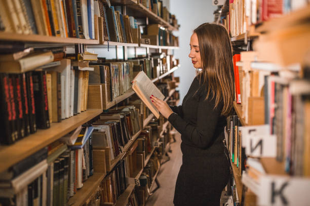 istock Side view of cheerful female student reading a book while standing leaning against bookshelf in library 2202829775