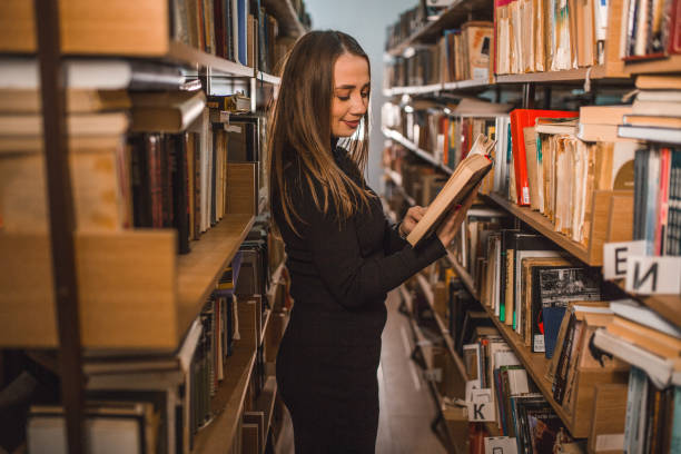 istock Side view of female student reading a book while standing leaning against bookshelf in library 2202829919