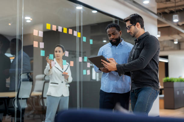 istock Diverse business professionals engaged in a discussion. A manager with a tablet leads the conversation while a colleague in the background places sticky notes on a glass wall, symbolizing teamwork, brainstorming, planning, and corporate success. 2202836224