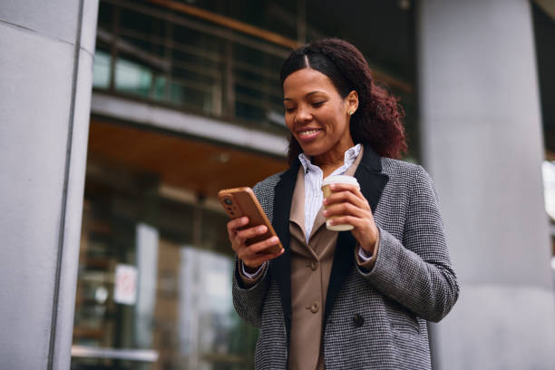istock Smiling businesswoman using smartphone and drinking coffee outside office building 2202842913