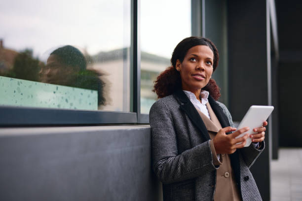 istock Businesswoman using digital tablet and looking away while leaning on office building 2202842959