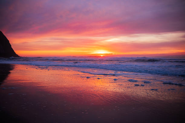 Ocean waves gently roll onto a dark sandy beach under a mesmerizing sunset. The sky is painted in warm shades of orange, pink, and purple, reflecting on the wet sand. A serene and relaxing atmosphere, perfect for concepts of travel, nature, and tranquility