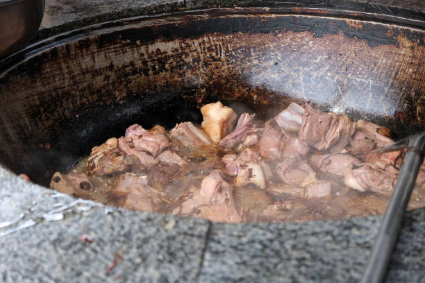 In a bustling outdoor kitchen, chunks of meat simmer in a large, round pot. Steam rises as flavors meld together. Local chefs are busy preparing traditional dishes for visitors