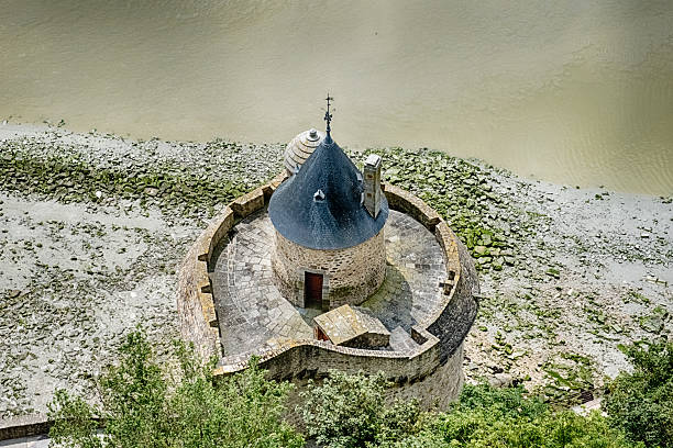 A small medieval stone house perched on a rocky outcrop at Mont Saint-Michel Castle in France. Photo taken at low tide. It's evening, the sky is blue and cloudy.