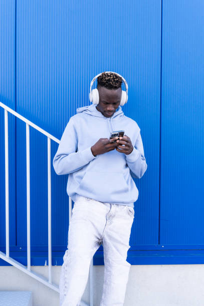 istock Young man wearing headphones and using a smartphone in front of a blue wall 2202877619