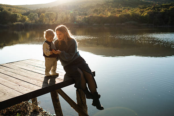 Stylish woman holding baby girl 2-3 year old wearing knitted sweaters sitting on wooden pier over lake and sunset outdoor. Motherhood.