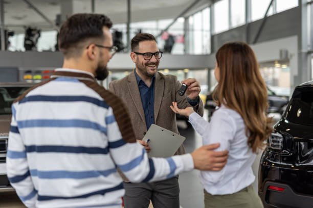 Happy car salesman giving keys to a couple in a dealership Smiling car salesman handing over keys to an excited young couple in a modern dealership, celebrating their new car purchase and marking a significant achievement in their lives car finance stock pictures, royalty-free photos & images