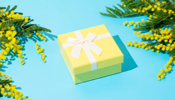 istock Yellow gift box on blue background with mimosa flowers and sunlight. 2202884352