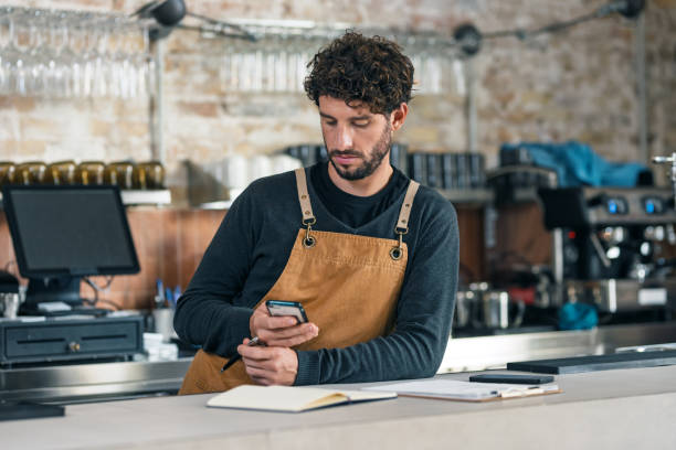 istock Handsome waiter taking inventory of everything missing while making a checklist on his phone at the coffee shop 2202888450