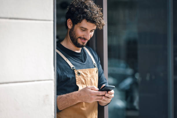 istock Handsome waiter using smartphone while leaning on the coffee shop door 2202888816