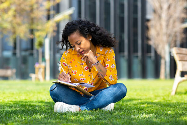 istock Young mixed race woman sitting on grass in park writing in book 2202920732