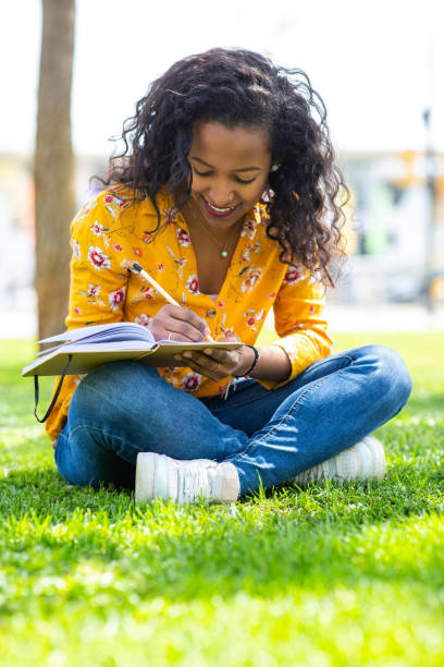 istock Young african woman sitting on grass in park writing in book 2202920733