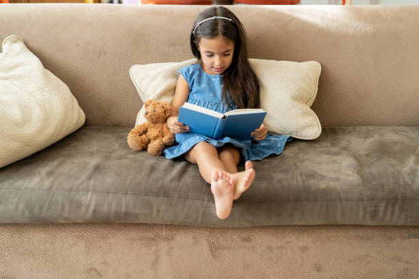 A small pretty girl is sitting on a couch with her teddy bear while she reads a fairy tale from a large book