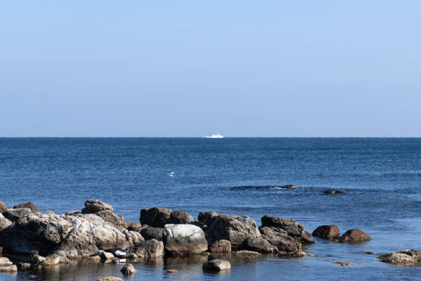 A serene shallow shoreline with scattered rocks, calm reflective water, and a soft blue sky streaked with clouds, evoking peaceful minimalism.