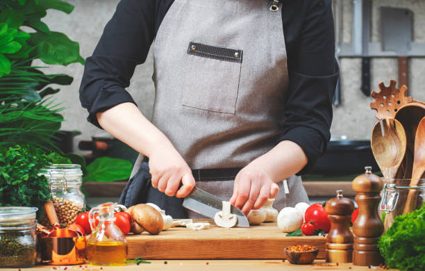 istock Chef cuts white champignon mushrooms. Cozy kitchen with wooden table, kitchenware, vegetables, herbs and ingredients for cooking. Healthy vegan food, culinary, recipes, food blogging 2202967020
